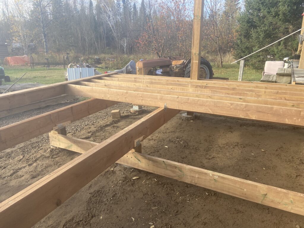 Deck floor joist framing on posts against a blue-sided Northern Ontario home.