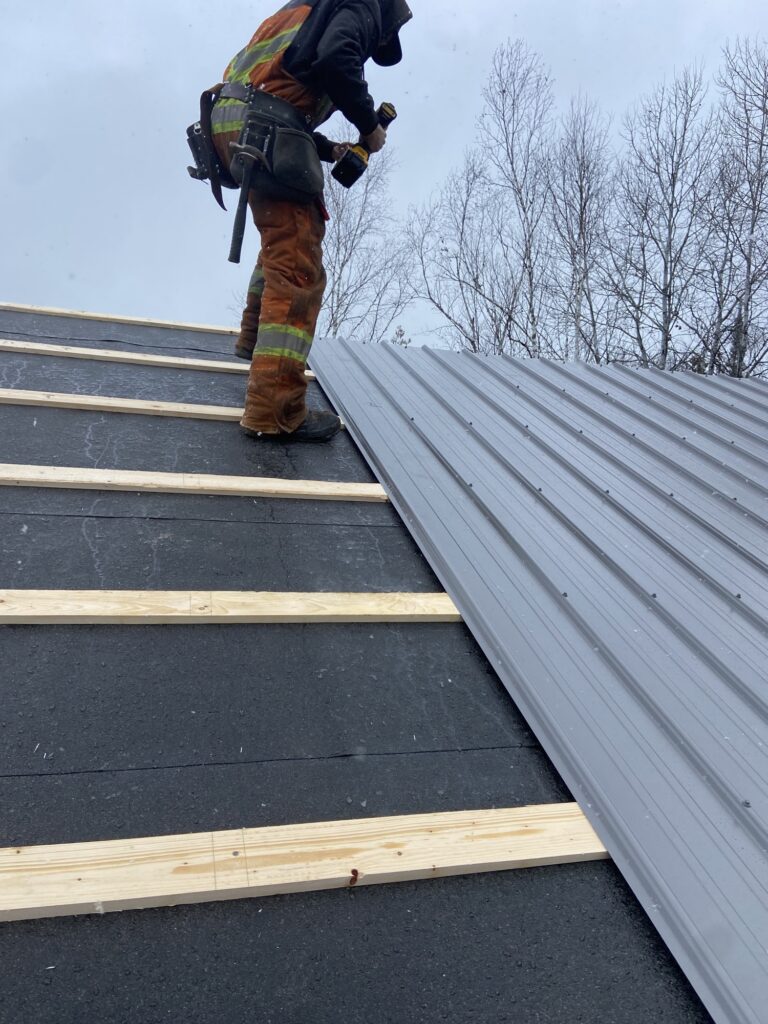 Crew installing grey metal roofing panels over underlayment and strapping.