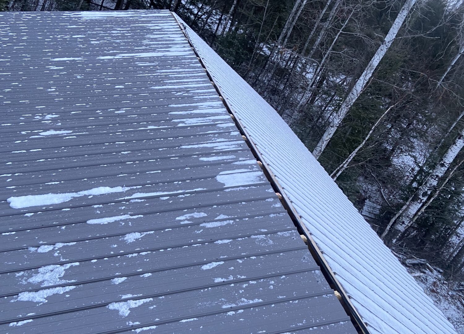 Finished grey metal garage roof dusted with the first snow, forest behind.