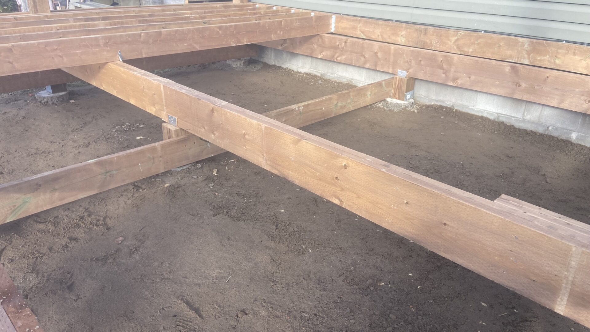 Pressure-treated deck beams and joists being framed over a leveled gravel base beside the house