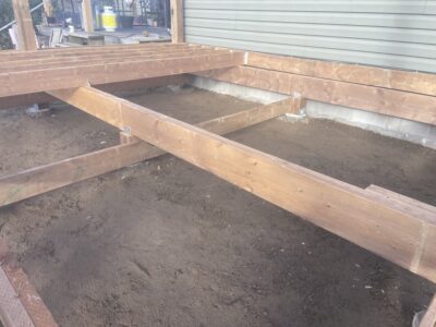 Pressure-treated deck beams and joists being framed over a leveled gravel base beside the house