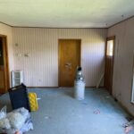 Room before renovation with striped wallpaper, a drop ceiling, and dark wood trim being cleared out