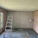 Room mid-demolition with peeling wallpaper and a stepladder beside the painted brick wall